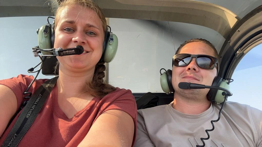 Twee personen met headsets zitten lachend in de cockpit van een helikopter.