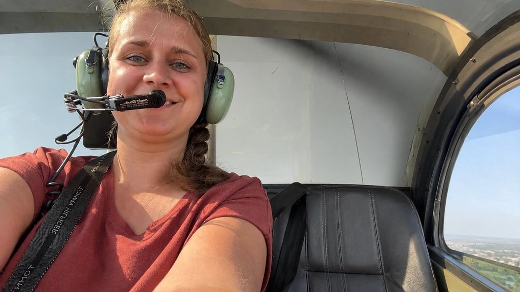 Een vrouw met een headset zit lachend in een helikopter, met naast haar een lege stoel en uitzicht door het raam op de lucht en het landschap.